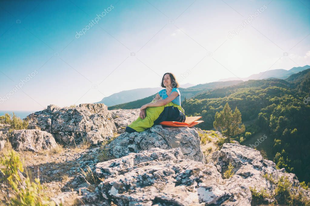 A woman is sitting in a sleeping bag at sunset. Camping on top of a mountain. A smiling girl rests after a climb. The brunette sits on a self inflating mat on the background of the mountains.
