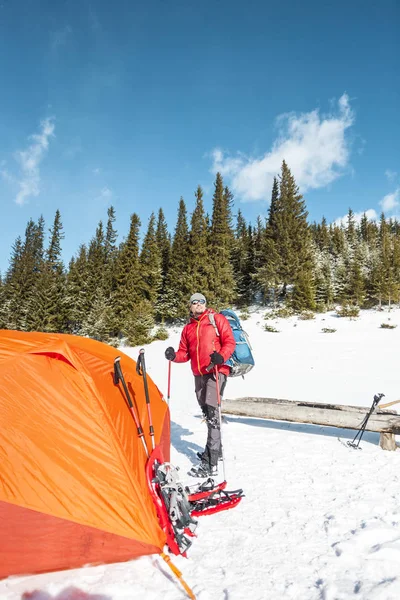 Man near the tent in winter. Tourist in extreme conditions. Snow shoes, trekking poles and backpack. Camping in the winter mountains. Orange tent in the snow.