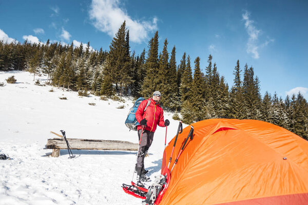 Man near the tent in winter. Tourist in extreme conditions. Snow shoes, trekking poles and backpack. Camping in the winter mountains. Orange tent in the snow.