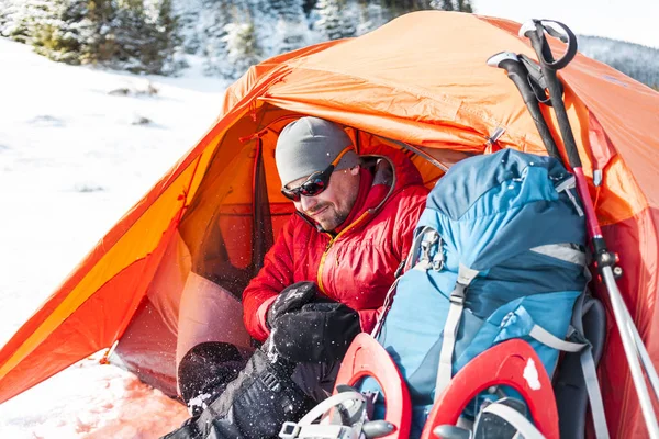 雪の中でキャンプ 冬の山でのトレッキングします オレンジ色のテント スノーシュー トレッキング ポール バックパックを持つ男は 雪の中のテントを設定します 極端な旅 冬のハイキングのための機器 — ストック写真