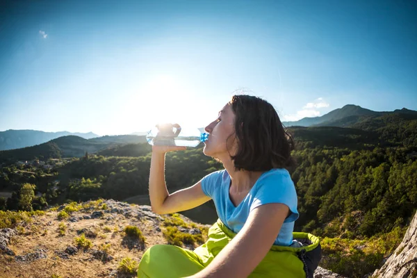 A woman is drinking water at the top of a mountain. The girl quenches his thirst. Tired tourist drinks from a plastic bottle. The brunette is sitting in a sleeping bag.