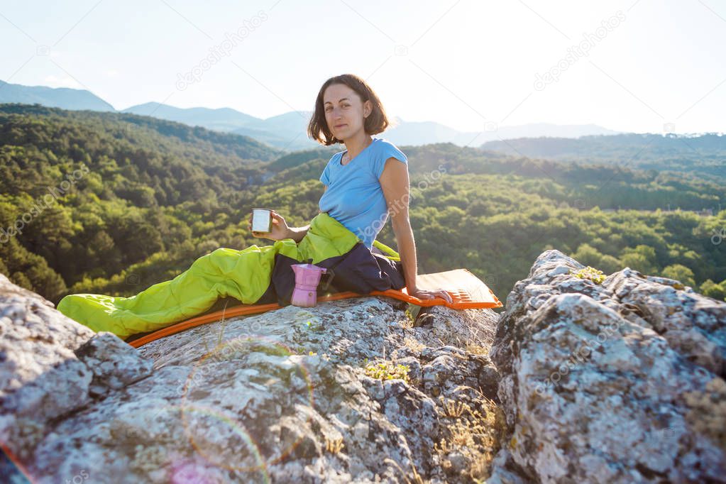 A woman is drinking coffee while sitting on top of a mountain. A girl in a sleeping bag drinks a hot drink from a mug. Smiling brunette. The traveler meets the dawn. Camping in nature.