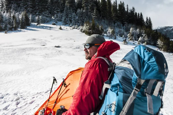 雪の中でキャンプ 冬の山でのトレッキングします オレンジ色のテント スノーシュー トレッキング ポール バックパックを持つ男は 雪の中のテントを設定します 極端な旅 冬のハイキングのための機器 — ストック写真