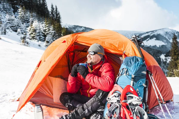 A man drinks from a mug near the tent in the winter. Tent, backpack, tracking sticks and snowshoes on the background of the mountains. Camping in the snow. Active rest and mountain climbing in winter.