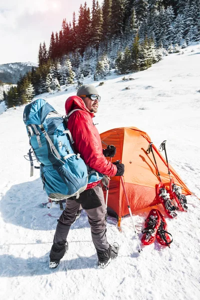 Man near the tent in winter. Tourist in extreme conditions. Snow shoes, trekking poles and backpack. Camping in the winter mountains. Orange tent in the snow.