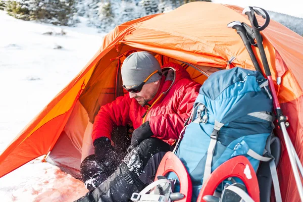 Camping in the snow. Winter trekking in the mountains. Orange tent, snowshoes and trekking poles. A man with a backpack sets a tent on the snow. Extreme journey. Equipment for winter hiking.