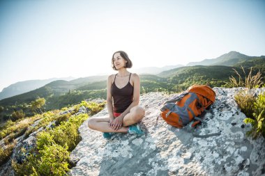 Sırt çantası bir kızla günbatımında bir dağın tepesinde oturuyor. Bir kadın yoga devreye girer. Meditasyon medeniyet çok uzak olduğunu. Güzel doğa ve tefekkür. Dağları nın görünümü.