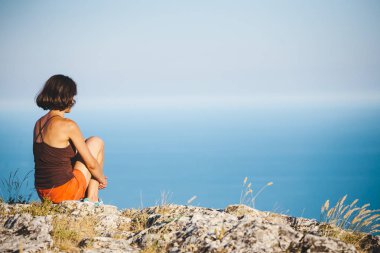 Bir kız günbatımında bir dağın tepesinde oturuyor. Bir kadın yoga yapan ve lotus pozisyonda oturuyor. Meditasyon medeniyet çok uzak olduğunu. Güzel kadın geri. Dağlar ve deniz görünümü.