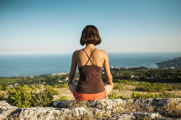 A girl is sitting on top of a mountain at sunset. A woman is engaged in yoga and sits in a lotus position. Meditation is far from civilization. Beautiful female back. View of the mountains and sea.