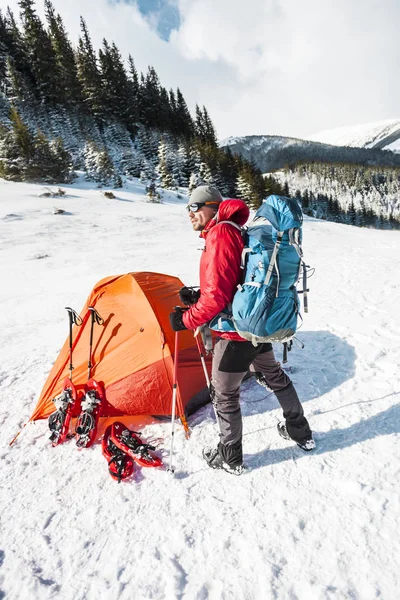 雪の中でキャンプ 冬の山でのトレッキングします オレンジ色のテント スノーシュー トレッキング ポール バックパックを持つ男は 雪の中のテントを設定します 極端な旅 冬のハイキングのための機器 — ストック写真