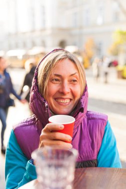 Bir kadın bir tek kullanımlık kağıt bardak kahve içiyor. Kız sıcak bir içecek bir şehir sokak içecekler. Gülümseyen sarışın.