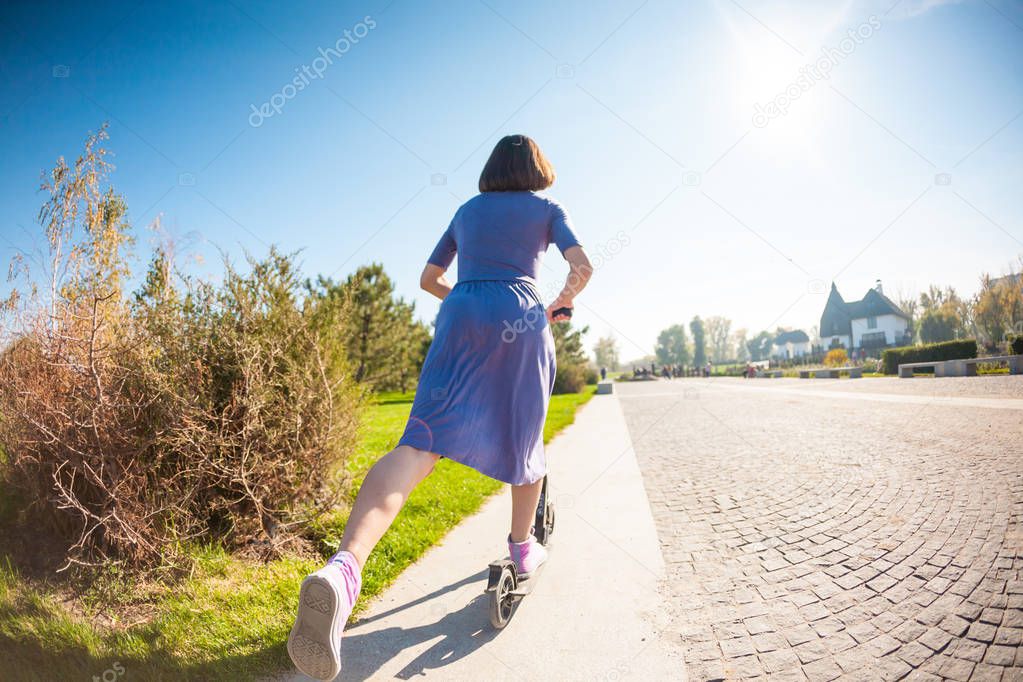 Slender girl in a dress and sneakers rides a scooter on the sidewalk. The woman put her foot to push away from the ground.