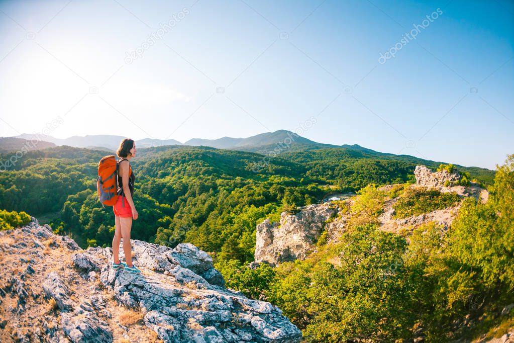 Girl at the top of the mountain. A woman with a backpack is standing on a rock. Climb to the top. Travel to picturesque places. Tourist against the sky and the sea. The brunette looks at the horizon.