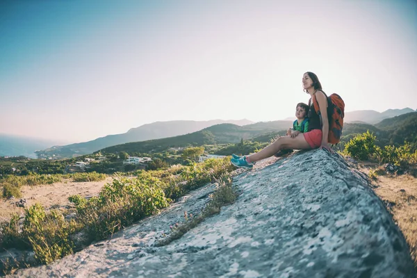 A woman is traveling with a child. Mother with a son is sitting on a large rock. Climb to the top of the mountain with children. The boy with the backpack climbed to the top. Active vacations.