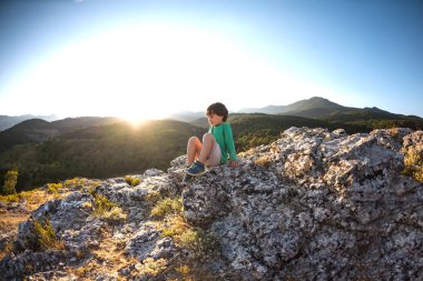 Gün batımında dağın zirvesine çocuğa. Çocuk zirveye tırmandı. Aktif tatiller. Çocuklarla seyahat. Çocuk bir kaya üzerinde oturuyor.