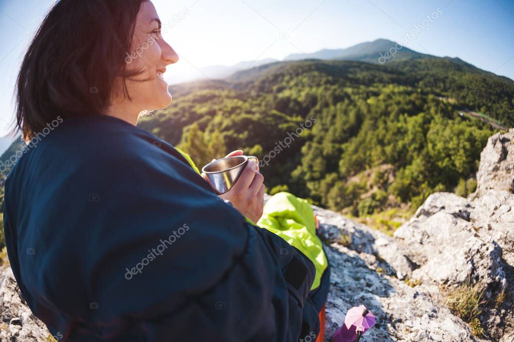 A woman is drinking coffee while sitting on top of a mountain. A girl in a sleeping bag drinks a hot drink from a mug. Smiling brunette. The traveler meets the dawn. Camping in nature. Fisheye lens.