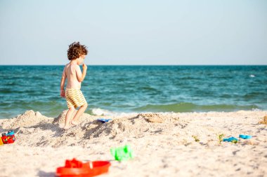 Bir çocuk bir deniz sahilde kum ile oynuyor. Çocuk gökyüzüne karşı kıyı boyunca çalışır. Çocuk doğada eğleniyor. Tatile çocuktur.