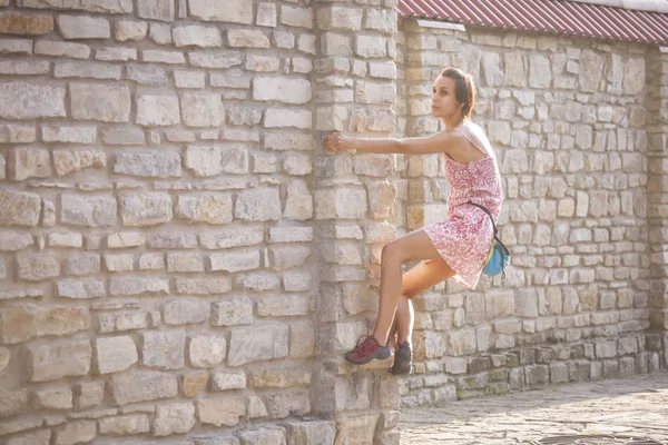 The girl climbs the stone wall. A woman in a summer dress climbs the wall of an old destroyed building. Brick fence. The climber is hanging on the city building. Strengthening the ruined castle.