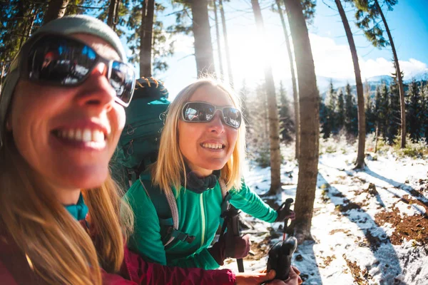 Two smiling women in a winter hike. Girlfriends with trekking poles are on a snow covered mountain path. Girls with backpacks and snow shoes travel together. Friends walk through the fir forest.