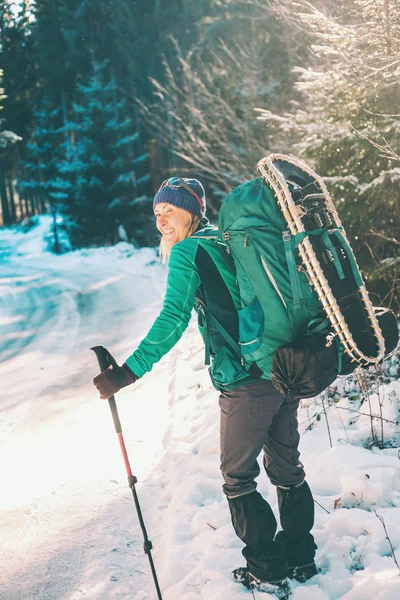 在冬天的山上背着背包和雪鞋的女人去风景秀丽的地方旅行 一个带着手杖的金发美女的画像 一个游客穿过白雪覆盖的森林 女孩沿着小路走着 — 图库照片
