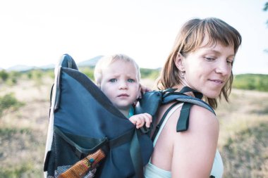Bir oğlan bir sırt çantası içinde oturuyor. Çocuk ailesiyle birlikte dolaşıyor. Bir çocukla tırmanma. Anne Bebek taşır. Küçük bir seyahat. Bir kadın bebeğini taşıyor. Çocuklarla seyahat.