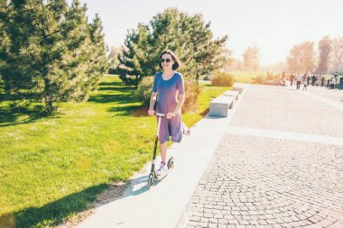 Kadın bir scooter sürme. Elbiseli kız parkta bir scooter sürmek. Esmer yolun dışına iter. Spor ayakkabı ayaklarda.