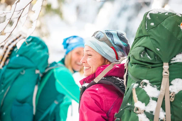两个微笑的女人在冬季远足 带着远足杆子的女朋友们在雪地覆盖的山路上 背着背包和雪鞋的女孩一起旅行 朋友们走过冷杉林 — 图库照片