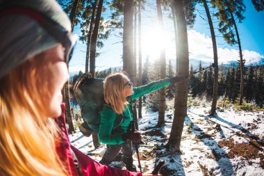 Kış yürüyüşünde gülümseyen iki kadın. Yürüyüş direkleri olan kızlar karla kaplı bir dağ yolundalar. Sırt çantalı ve kar ayakkabılı kızlar birlikte seyahat eder. Dostlar köknar ormanında yürür.