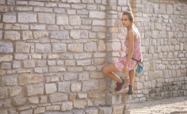 Bir kadın elbiseli bir tuğla duvara tırmanıyor. Kız taş bir çite tırmanıyor. Tırmanıcı bir şehir sokak üzerinde. Eski yıkılan binanın duvarına.