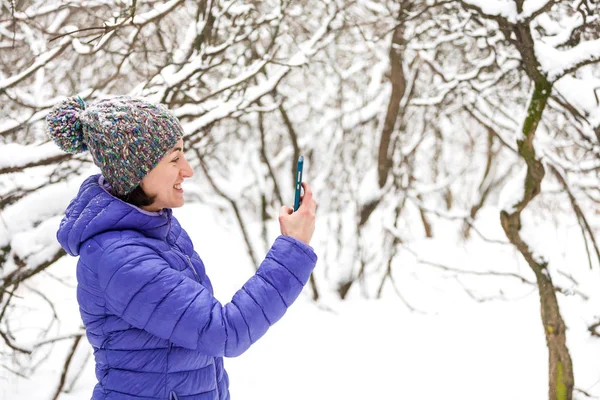 Smartphone screen. A woman makes a photo on a mobile phone in the winter forest. A smiling girl in a knitted hat makes selfie against the background of a snow covered park.