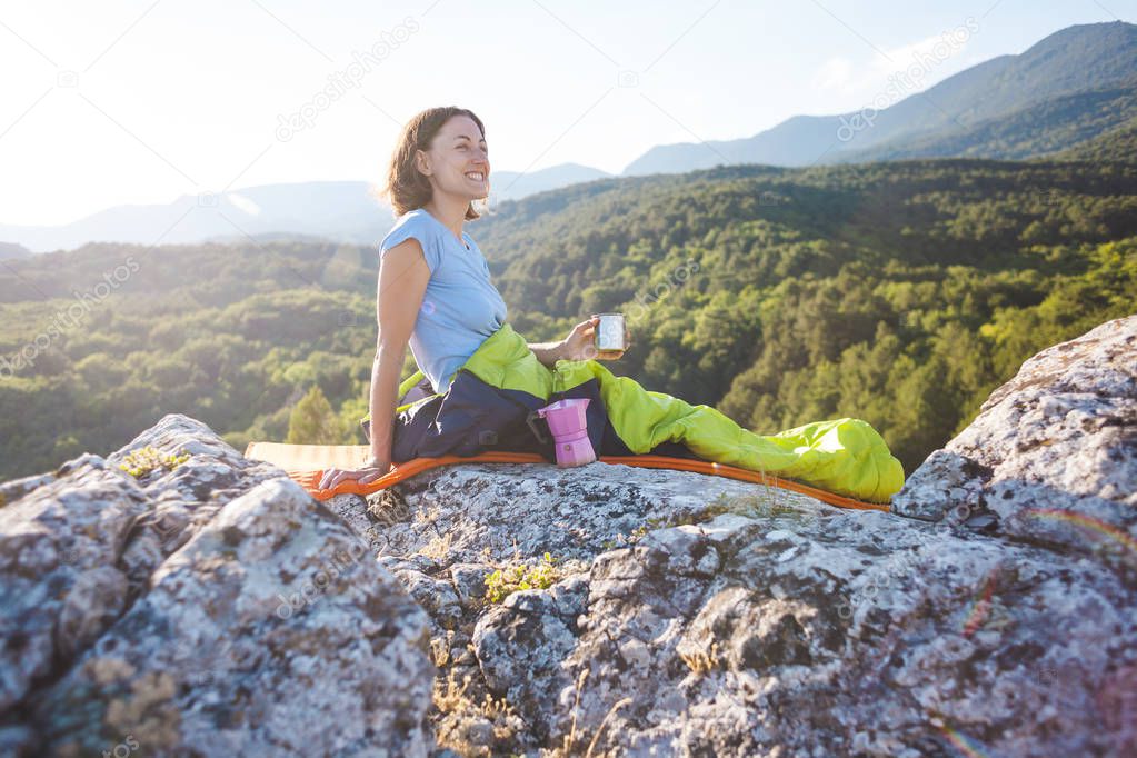 A woman is drinking coffee while sitting on top of a mountain. A girl in a sleeping bag drinks a hot drink from a mug. Smiling brunette. The traveler meets the dawn. Camping in nature.
