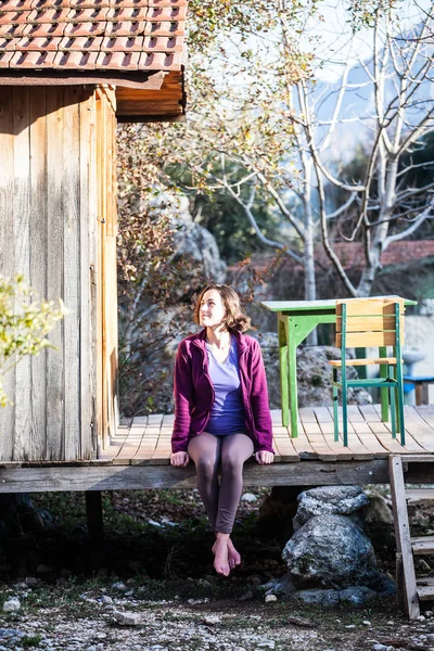 A woman is sitting on the porch of an old wooden house. Barefoot girl resting near the house on a warm spring day.