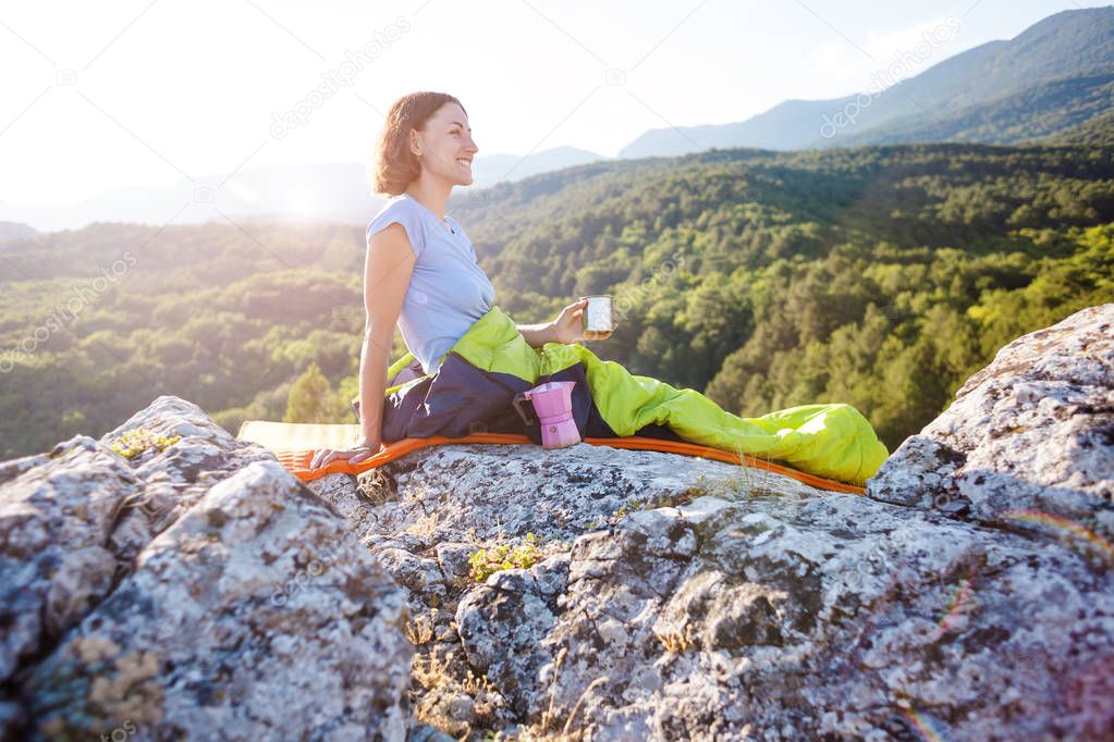 A traveler is drinking coffee while sitting on top of a mountain. A girl in a sleeping bag drinks a hot drink from a mug. Smiling brunette. The woman meets the dawn. Camping in nature.