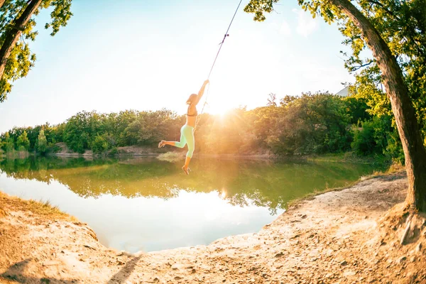 Jump into the water. A woman is resting on the lake. A swing from a rope and a stick. Active recreation in nature. Summer fun. A woman is riding a swing. Fisheye lens.