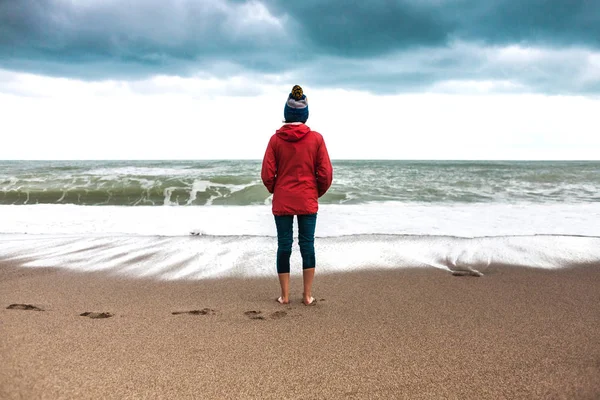 Barefoot woman looks at the winter sea. The girl in the cap walks along the coast of the ocean on a cloudy day. A woman wets her feet in cold water. Waves and sky on an autumn day.