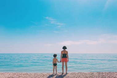 Annesi sahilde duran ve denize bakan bir çocuk.