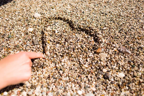 Uma menina senta-se na praia e desenha um coração na areia . — Fotografia de Stock