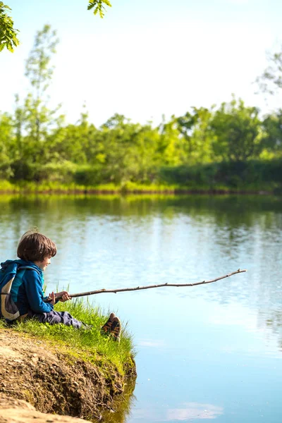 Хлопчик з рюкзаком сидить на озері з дерев'яною паличкою . — стокове фото