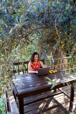 Kırmızı elbiseli bir kadın ahşap bir evin terasında çalışıyor, bir kız dizüstü bilgisayara bakıyor, uzaktan eğitim görüyor, serbest çalışan biri evin verandasında çalışıyor, karantina sırasında uzaktan kumandalı iş yapıyor..