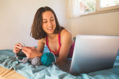 Bir kadın internetten video dersleri öğreniyor, yeni beceriler geliştiriyor, örgü örüyor, kız kanepede oturup dizüstü bilgisayar izlerken örgü örüyor, karantina sırasında hobiler ediniyor..