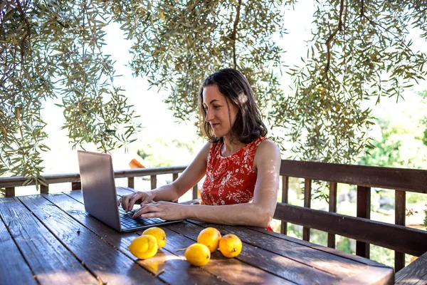 A woman in a red dress works on the terrace of a wooden house, A girl looks into a laptop, Distance education, Freelancer works on the veranda of the house, Remote work during quarantine.