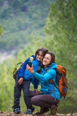 Sırt çantalı bir çocuk, bir dağ ormanının arka planında annesiyle birlikte akıllı telefondan selfie çeker, bir çocuk annesiyle seyahat eder, bir kadın oğluyla fotoğraf çekilir, çocuklarla yürüyüş yapar..