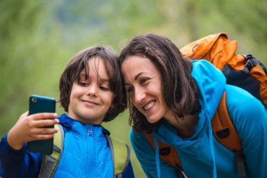 Sırt çantalı bir çocuk, bir dağ ormanının arka planında annesiyle birlikte akıllı telefondan selfie çeker, bir çocuk annesiyle seyahat eder, bir kadın oğluyla fotoğraf çekilir, çocuklarla yürüyüş yapar..