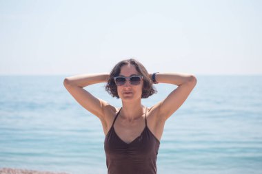 Gülümseyen bir kadın kıyıda duruyor. İnce kız okyanusta dinleniyor. Kumsalda elleri başının arkasında duran esmer bir kadın..