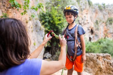 Eğitmen çocuğa tırmanma ekipmanlarını kullanmayı öğretiyor, çocuk kayaya tırmanmaya hazırlanıyor, bir kadın bir çocuğa bağlanmak için karabinayı nasıl kullanacağını gösteriyor, anne ipi güvenlik sistemine bağlıyor..