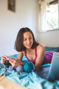 Bir kadın internetten video dersleri öğreniyor, yeni beceriler geliştiriyor, örgü örüyor, kız kanepede oturup dizüstü bilgisayar izlerken örgü örüyor, karantina sırasında hobiler ediniyor..