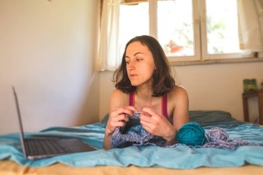 Bir kadın internetten video dersleri öğreniyor, yeni beceriler geliştiriyor, örgü örüyor, kız kanepede oturup dizüstü bilgisayar izlerken örgü örüyor, karantina sırasında hobiler ediniyor..