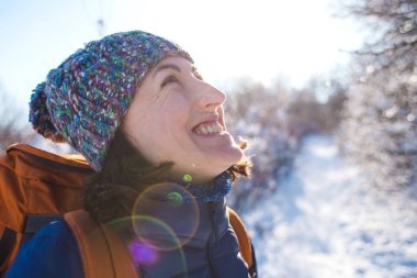 Örgü şapkalı bir kızın portresi. Karlı bir ormanın arka planında sırt çantalı gülümseyen bir kadın. Kış yürüyüşüne çıkmış bir kadın. Kış yükseliyor dağlara.