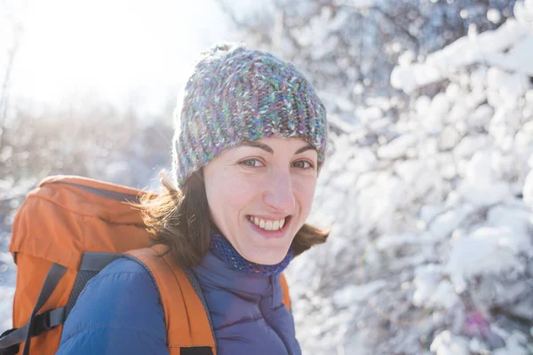 Portrait of a girl in a knitted hat. Smiling woman with a backpack on a background of a snowy forest. Woman on a winter hike. Winter ascent to the mountains.