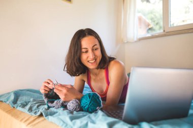 Bir kadın internetten video dersleri öğreniyor, yeni beceriler geliştiriyor, örgü örüyor, kız kanepede oturup dizüstü bilgisayar izlerken örgü örüyor, karantina sırasında hobiler ediniyor..